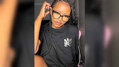 Media: Video of a Black woman with braided hair, wearing black glasses, a black t-shirt with white text, and pink jacket, seated indoors.