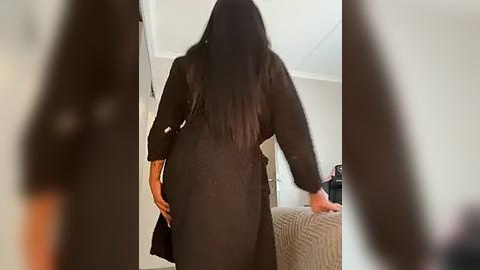 Media: Video of a woman with long black hair, wearing a black robe, standing in a bedroom with beige furniture.