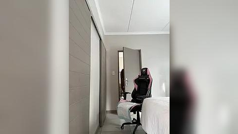 Media: Video of a minimalist, beige-tiled bathroom with a closed door, open door to the right, and a gaming chair positioned in the doorway.