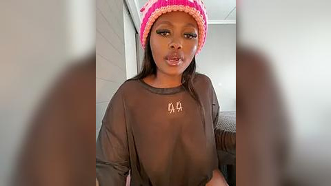 Media: A video of a young black woman with medium brown skin, wearing a sheer black shirt, pink beanie, and black leggings. The background shows a dimly lit room with neutral-colored walls.