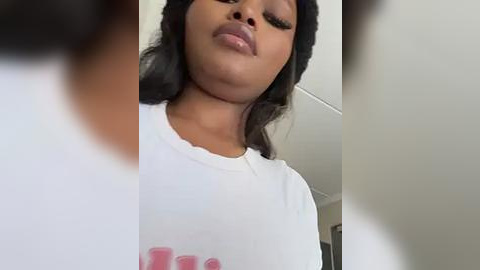 Media: Video of a young Black woman with medium brown skin, wearing a white T-shirt, long black hair, and full lips, indoors with blurred background.