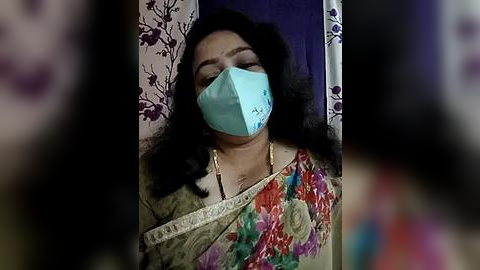 Media: Video of a woman in a floral saree, wearing a light blue surgical mask, sitting against a floral-patterned backdrop, blurred background, and indistinct people.