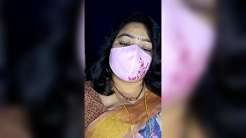 Media: Video of a South Asian woman with medium skin tone, wearing a pink mask, mustard-yellow saree, and a gold necklace, against a dark, blurred background.