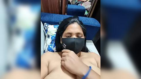 Media: A video of a topless woman with medium brown skin, black mask, and dark hair, lying on a blue couch in a cluttered room.