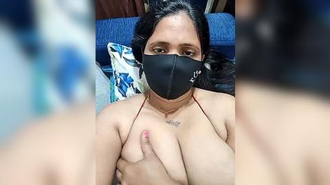 Media: A video of a topless South Asian woman with medium skin tone, black hair, and a black mask, covering her breasts with one hand, sitting on a blue cushioned couch.