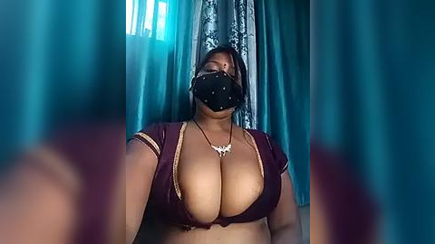 Media: Video of a woman with large breasts, wearing a maroon bra, black face mask, and a necklace, indoors with teal curtains.