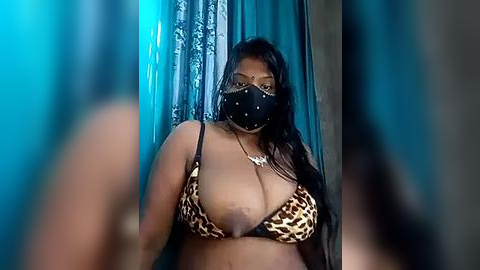 Media: Video of a dark-skinned woman with long black hair, wearing a leopard-print bra, black face mask, and a gold necklace, standing in front of teal curtains.