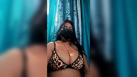 Media: Video of a curvy woman with dark hair and a large bust, wearing a leopard-print bra and a black face mask, standing in front of a teal curtain with a floral pattern.