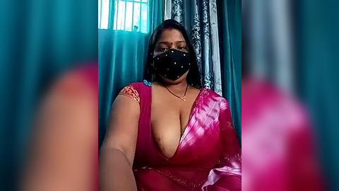 Media: A video shows a South Asian woman with medium skin tone, wearing a black face mask and a low-cut, pink satin dress, with a teal curtain backdrop and blurred foreground.