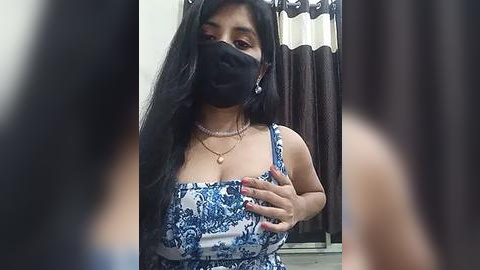 Media: Video of a South Asian woman with long black hair, wearing a blue floral-patterned dress, black mask, and a gold necklace, indoors, blurred background.