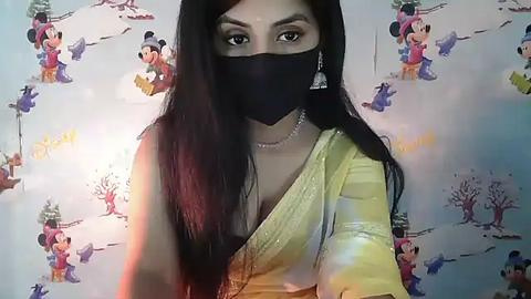 Media: A video of a South Asian woman with long black hair, wearing a black mask and a yellow saree, against a background of Disney characters in a snowy setting.