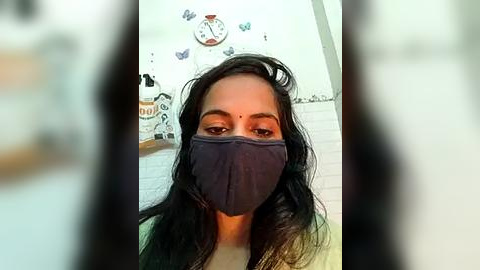 Media: Video of a South Asian woman with long black hair, wearing a blue mask, white top, and a white wall with a clock and butterfly stickers in the background.