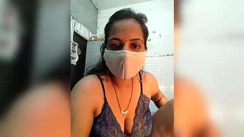 Media: Video of a South Asian woman with medium-dark skin, wearing a blue patterned camisole, white mask, and long black hair in a bun. Background includes a gray wall with a calendar, white window blinds, and a small table.