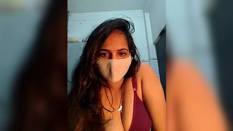 Media: Video of a woman with long dark hair, wearing a mask, red top, and necklace, indoors with blurred foreground.