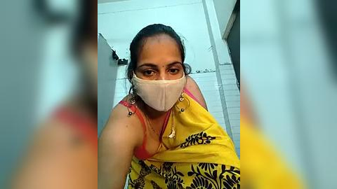 Media: Video of a woman with medium brown skin, wearing a yellow saree, red tank top, and a white mask, taken from a low angle, indoors, with blurred background.