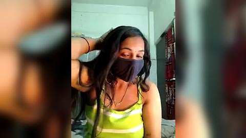 Media: Video of a young woman with long, dark hair, wearing a black face mask, green striped tank top, and a black bracelet, sitting in a dimly lit room with a red curtain and a white wall in the background.