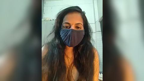 Media: Video of a young woman with long, dark hair, wearing a navy blue face mask, standing indoors with a blurred background.