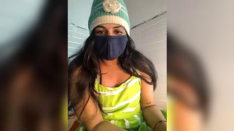 Media: Video of a young woman with long black hair, wearing a green and white striped top, a teal knit beanie, and a navy face mask. She sits on a bed with white blinds in the background.