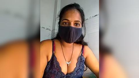 Media: Video of a South Asian woman with medium skin tone, wearing a navy blue lace bra and face mask, looking into the camera. Background shows a plain, white wall.