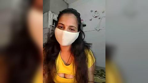 Media: Video of a young woman with dark hair, wearing a yellow shirt, a white face mask, and a bindi, indoors with a blurry background featuring a wall with sketches.