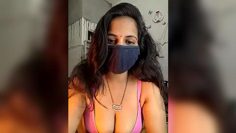 Media: Video of a South Asian woman with long, wavy black hair, wearing a pink tank top, blue face mask, and a gold necklace. Background shows a blurred room with white clock and wall art.