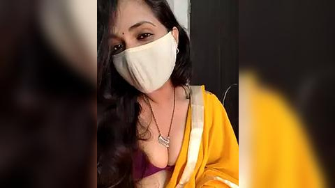 Media: Video of a woman with long, wavy black hair, wearing a beige face mask, a purple bra, and a yellow sari, with a blurred background.