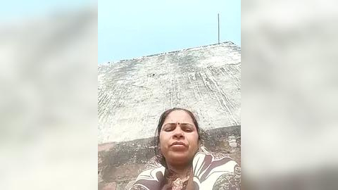 Media: Video of a smiling woman with dark hair wearing a striped shawl, standing in front of a crumbling, sunlit stone wall against a clear blue sky.