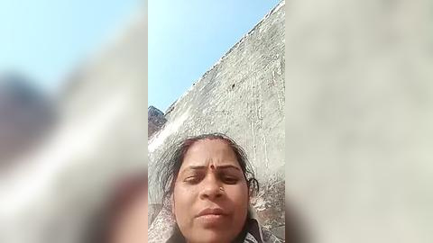 Media: A video of a young woman with a serene expression, her head framed by a large, cracked, white plaster wall, with blurred out-of-focus people in the foreground.