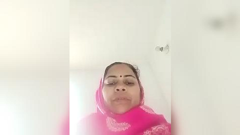 Media: A video of a woman in a bright pink saree with white floral patterns, wearing a red bindi, and a white headscarf. The background is a white wall and door, with a small, shiny metal object on the wall.