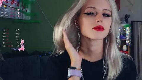 Media: Video of a young woman with platinum blonde hair, red lipstick, and dramatic winged eyeliner, wearing a black top, in a dimly lit room with a green wall and a TV displaying chat messages.