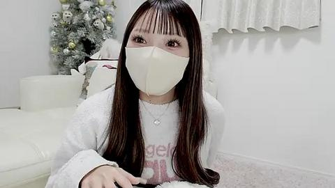 Media: Video of a young Asian woman with long, straight dark hair, wearing a beige mask and white sweater, sitting on a white carpet in a modern, minimalist room with a Christmas tree and white curtains.