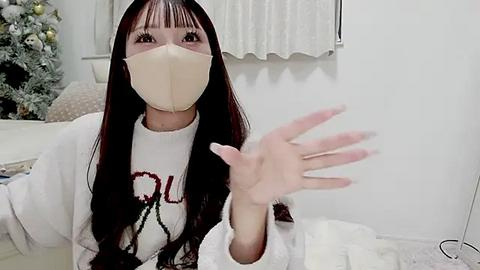 Media: Video of an East Asian woman with long black hair, wearing a beige mask, white sweater, and beige gloves, waving her hand in a modern, minimalist room with a snow-covered Christmas tree in the background.