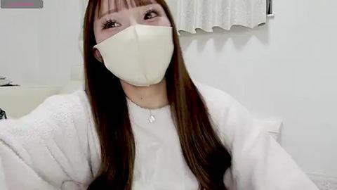 Media: Video of an Asian woman with long brown hair, wearing a white mask, white cardigan, and a necklace, seated in a brightly lit room with white walls and a window with white curtains.