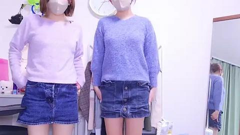 Media: Video of two young women with light skin, wearing light pink and blue denim outfits, standing in a small room with a mirror, pink stuffed animal, and a clock on the white wall.