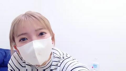 Media: Video of a young Asian woman with light skin and short blonde hair, wearing a white face mask, black-and-white striped shirt, and blue fabric. She looks into the camera with a neutral expression against a plain white background.