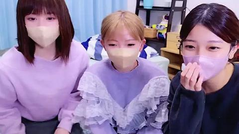 Media: Video of three East Asian women with light skin, wearing light purple and black sweaters, light masks, and straight brown hair, sitting closely together in a modern, softly lit room.