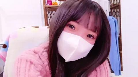 Media: Video of a young Asian woman with straight black hair, wearing a white face mask, pink fuzzy robe, and sitting in a cluttered room with a shelf of toys and clothes in the background.