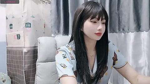 Media: Video of an East Asian woman with long black hair, fair skin, and red lipstick, wearing a light blue shirt with cartoon designs, sitting on a white couch with gray curtains in the background.