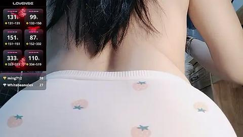 Media: Video of a person with light skin and long black hair, wearing white panties with strawberry patterns, captured from behind, revealing their bare back. The background is blurred, possibly a bedroom setting.