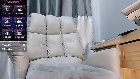 Media: Video of a plush, cream-colored, tufted leather armchair with a wooden side table beside it. A digital weather forecast overlay shows rain. The room has light blue walls and a gray curtain partially covering a window.