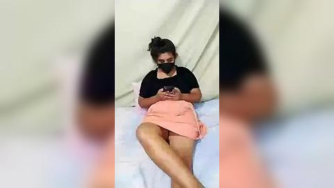 Media: Video of a woman lying on a bed in a hospital room, wearing a black mask and peach-colored dress. Her legs are crossed, and she holds a phone. The room is plain, with a beige curtain in the background.