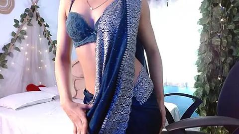 Media: Video of a slender, light-skinned woman with a small bust wearing a blue, lace-trimmed bra and a matching sari, standing in a bright room with a bed, green plants, and a balcony view.