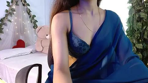 Media: A video of a South Asian woman with long, dark hair wearing a blue sari and a lace bra, sitting on a bed with a teddy bear, fairy lights, and green plants in the background.