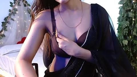 Media: Video of a young woman with long brown hair, wearing a sheer black blouse, partially revealing a black bra, in a softly lit bedroom with fairy lights and greenery.