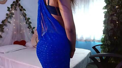 Media: Video of a woman in a sheer blue sari with a black bra, standing in a dimly-lit bedroom with a bed, fairy lights, and a window.