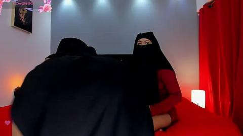 Media: Video of a woman with a black hijab lying on a red carpet, visible from the back, in a dimly lit room with red curtains and a lit lamp.