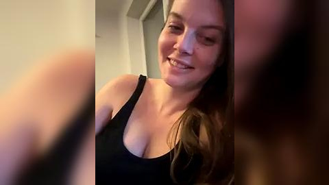 Media: Video of a smiling woman with long brown hair, wearing a black tank top, in a dimly lit room.