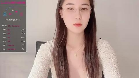 Media: Video of a young East Asian woman with long, straight black hair and fair skin, wearing wire-rimmed glasses and a white lace top, seated against a plain beige wall.