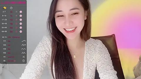 Media: Video of a smiling East Asian woman with long black hair, wearing a white lace cardigan, sitting on a dark brown chair. Background features a colorful, abstract wall painting.