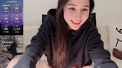 Media: A video of a smiling, light-skinned Asian woman with long, straight black hair, wearing a black hoodie, sitting on a white couch. The background shows a TV displaying weather information.